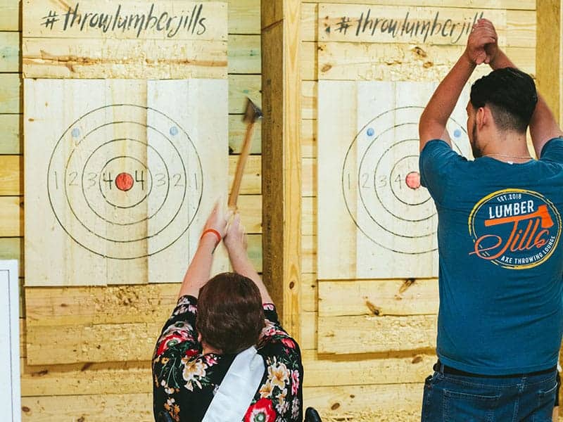 Image of Lumber Jill's employee teaching a PWD adult female on how to throw an axe to a target at Lumber Jill's axe throwing lane - Image of kids with adults enjoying their time at Lumber Jill's axe throwing lounge - Image of an adult male posing at his bull's eye target at Lumber Jill's axe throwing - www.throwlumberjills.com