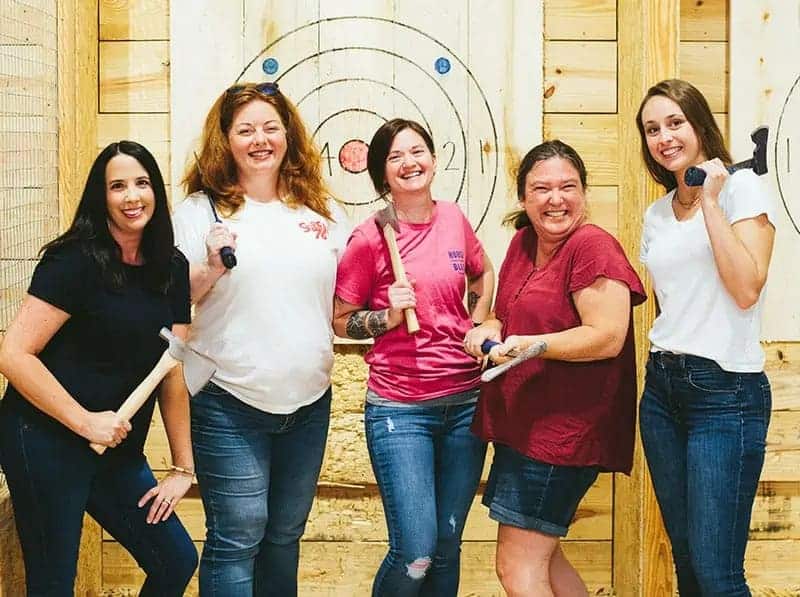 Image of 5 adult females each holding an axe posing for a photo at Lumber Jill's axe throwing lane target - Image of kids with adults enjoying their time at Lumber Jill's axe throwing lounge - Image of an adult male posing at his bull's eye target at Lumber Jill's axe throwing - www.throwlumberjills.com